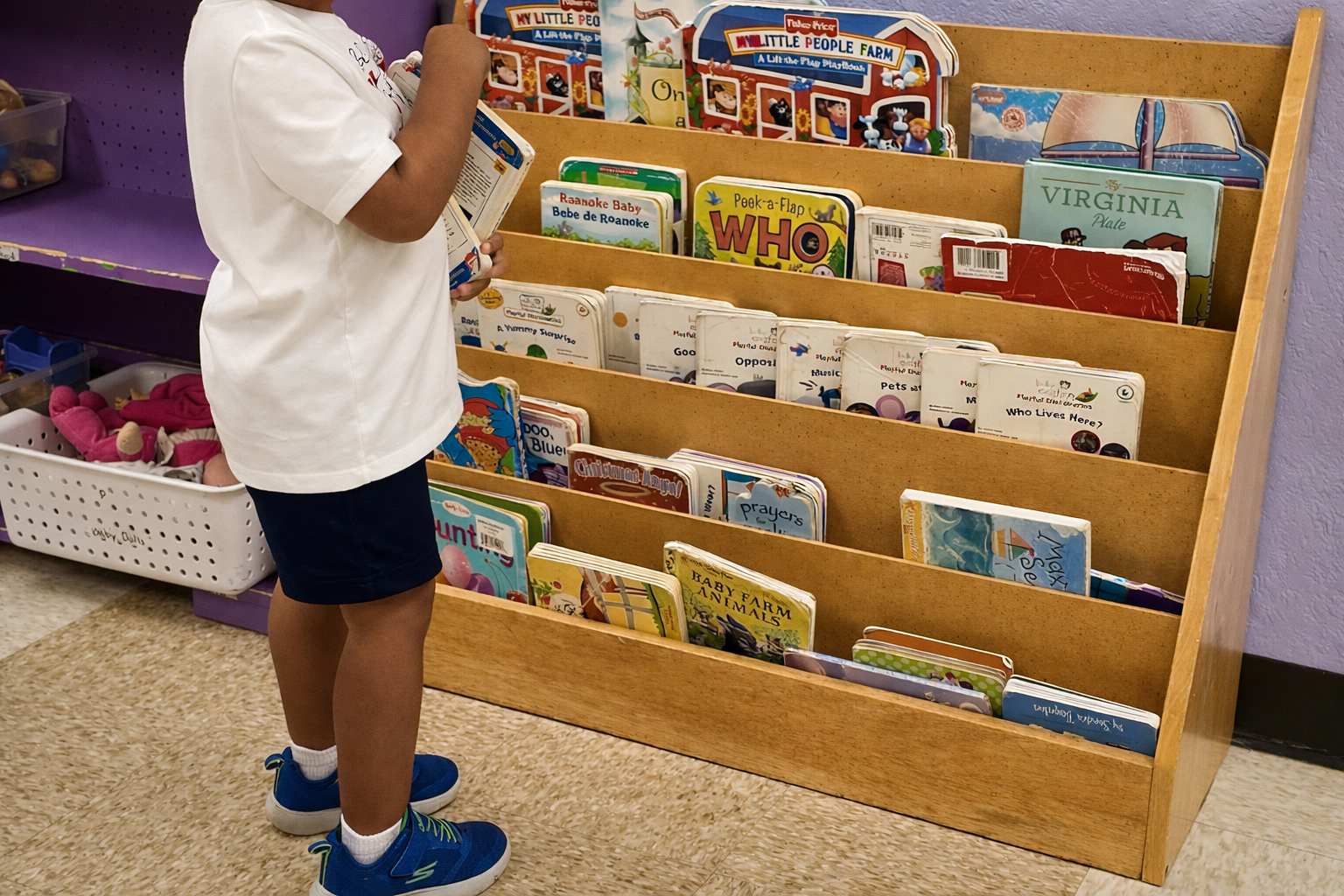 Child standing by a wooden bookshelf of colorful books in a classroom or library.