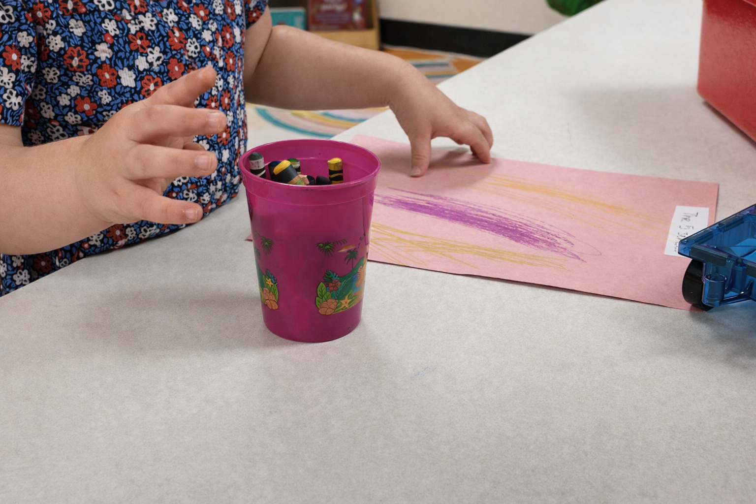 Child working on a pink craft paper beside a purple cup on a table