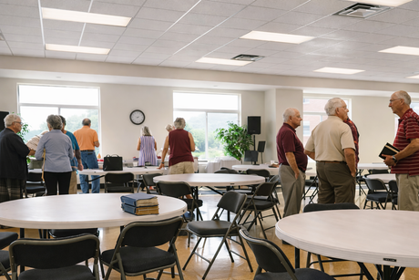 People standing and chatting in a bright fellowship hall with round tables and folding chairs