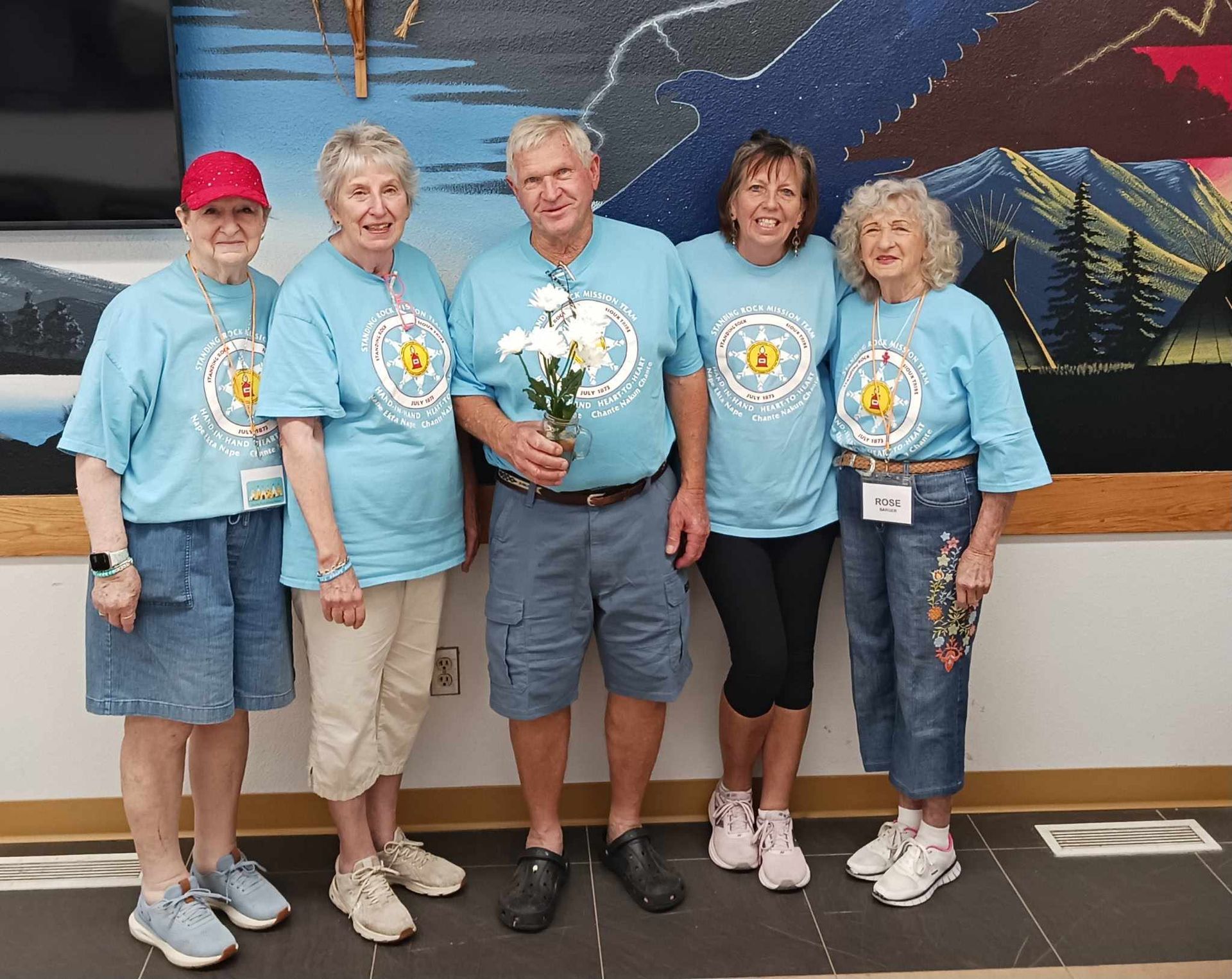 Five volunteers in matching blue shirts pose indoors, one holding flowers, in front of a mural.