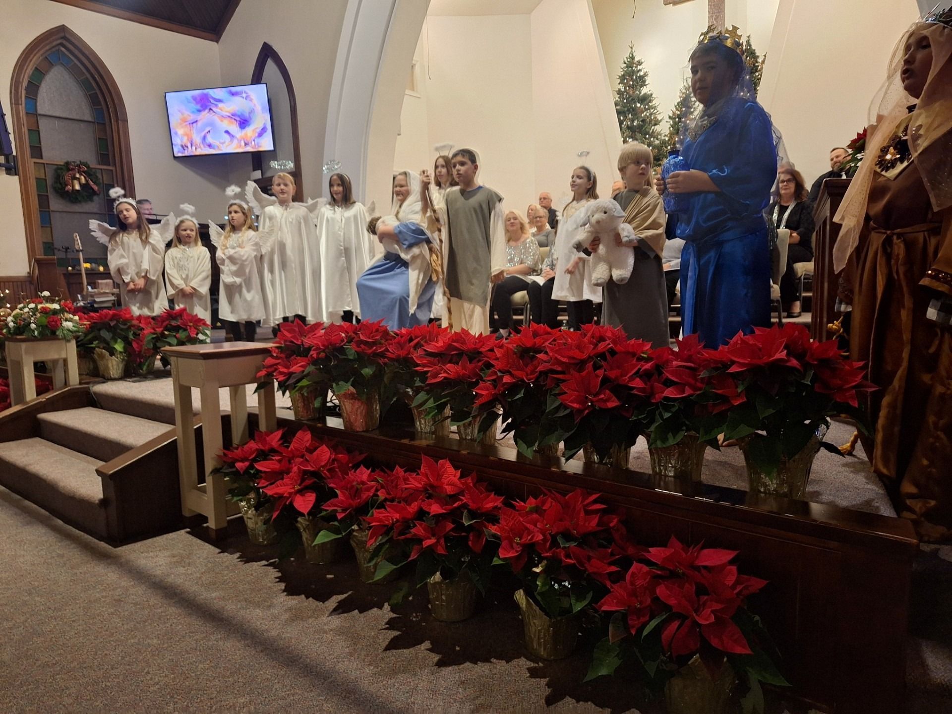 Children in white robes stand in a church, with red poinsettias in front.