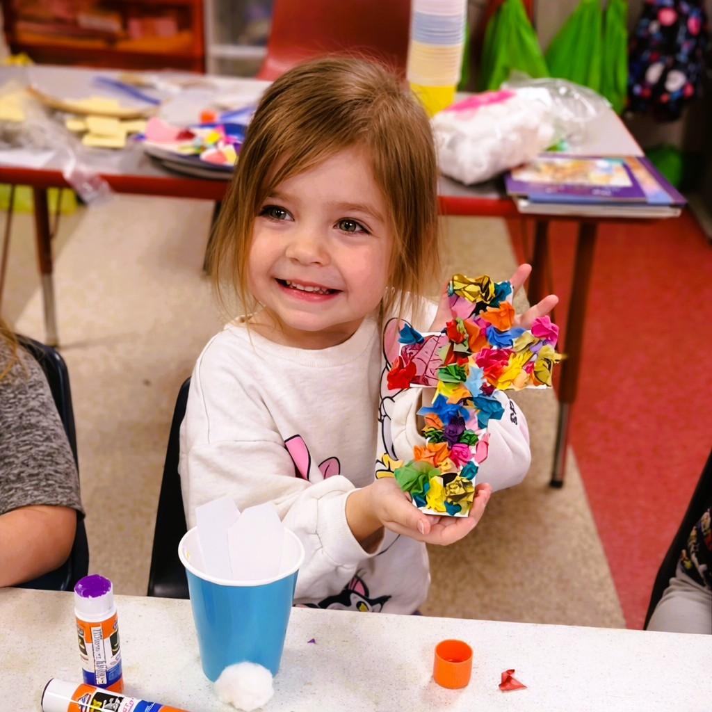 Child smiling while holding a colorful craft made from tissue paper at a classroom table.