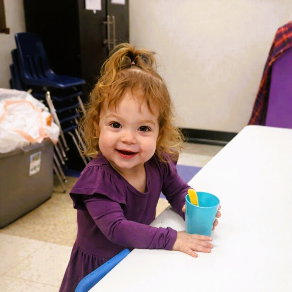 Smiling child in a purple dress holding a blue cup at a white table in a classroom