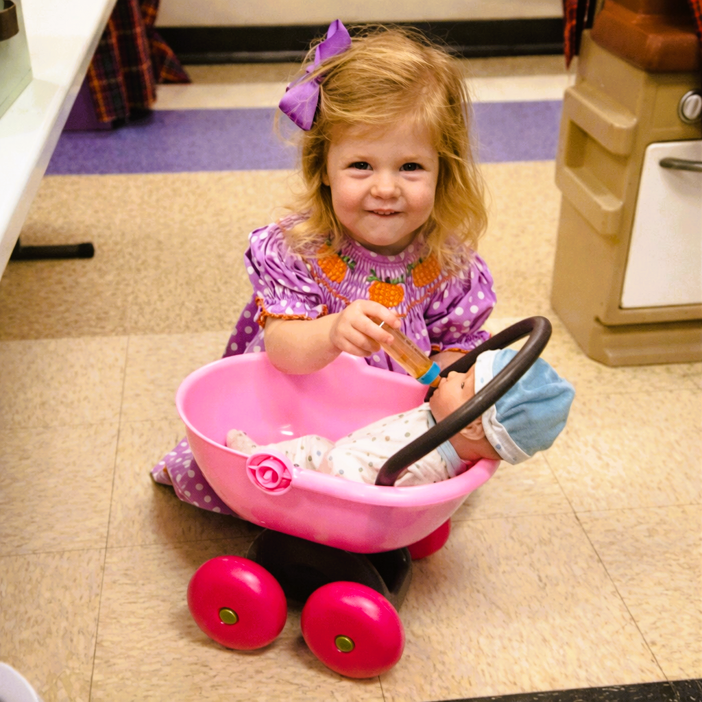 Toddler in a purple dress sits in a pink toy stroller, smiling and holding the handle indoors.