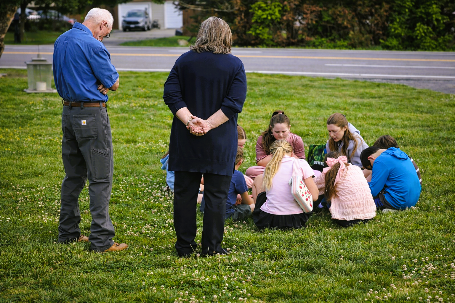 Small group sitting on grass outdoors while two adults stand nearby, one holding a clipboard.