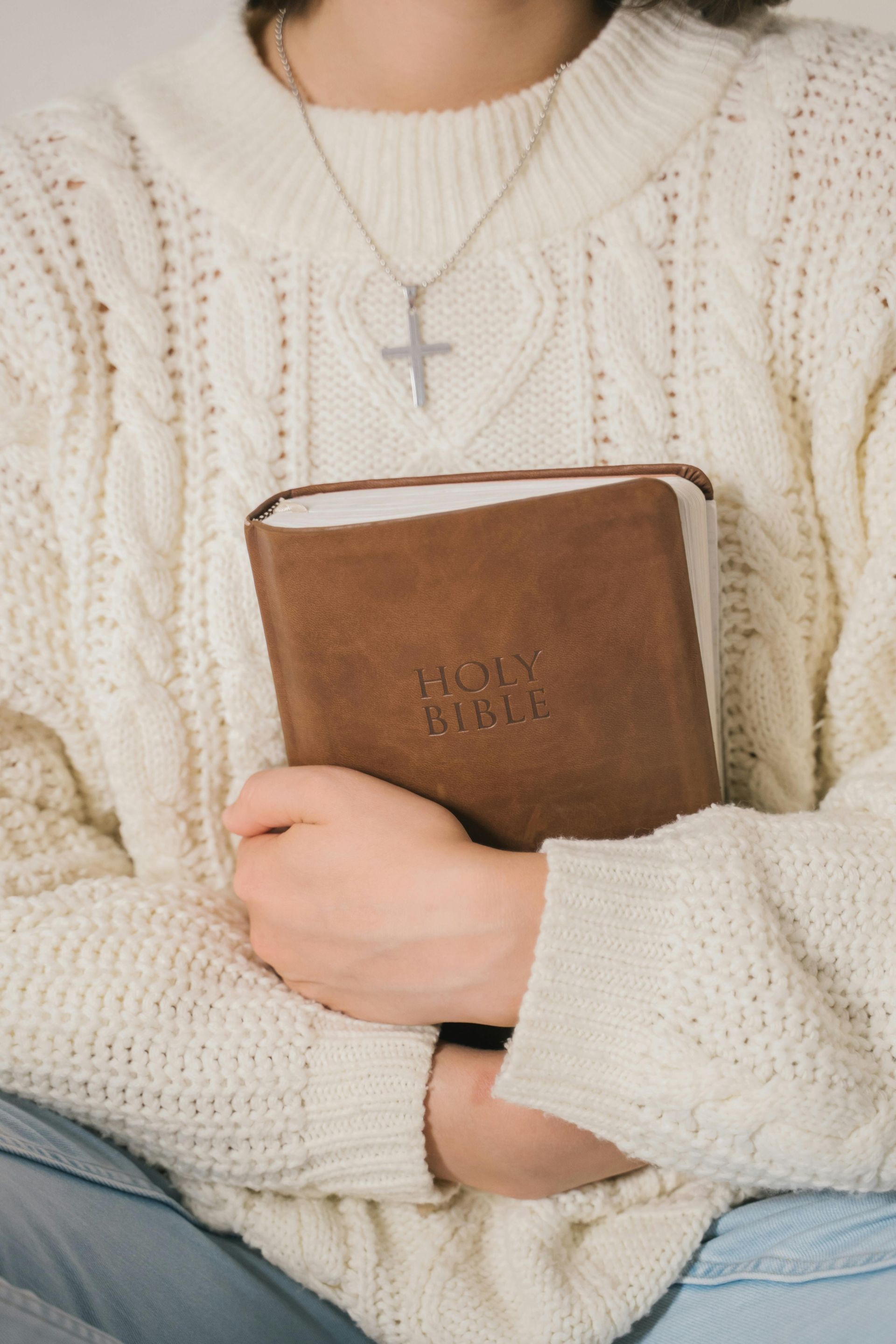 Person in cream sweater holding a brown Bible with a silver cross necklace visible