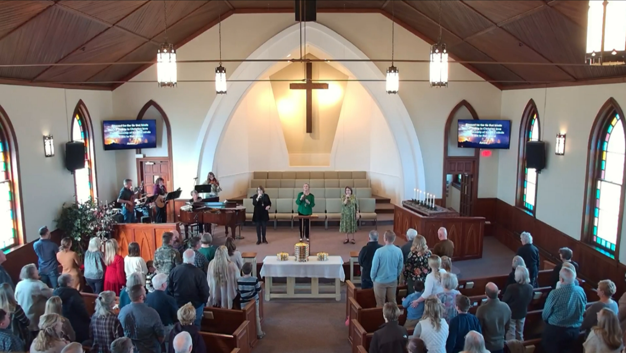 Congregation gathered in a church sanctuary around the altar, with a large cross and stained-glass windows.