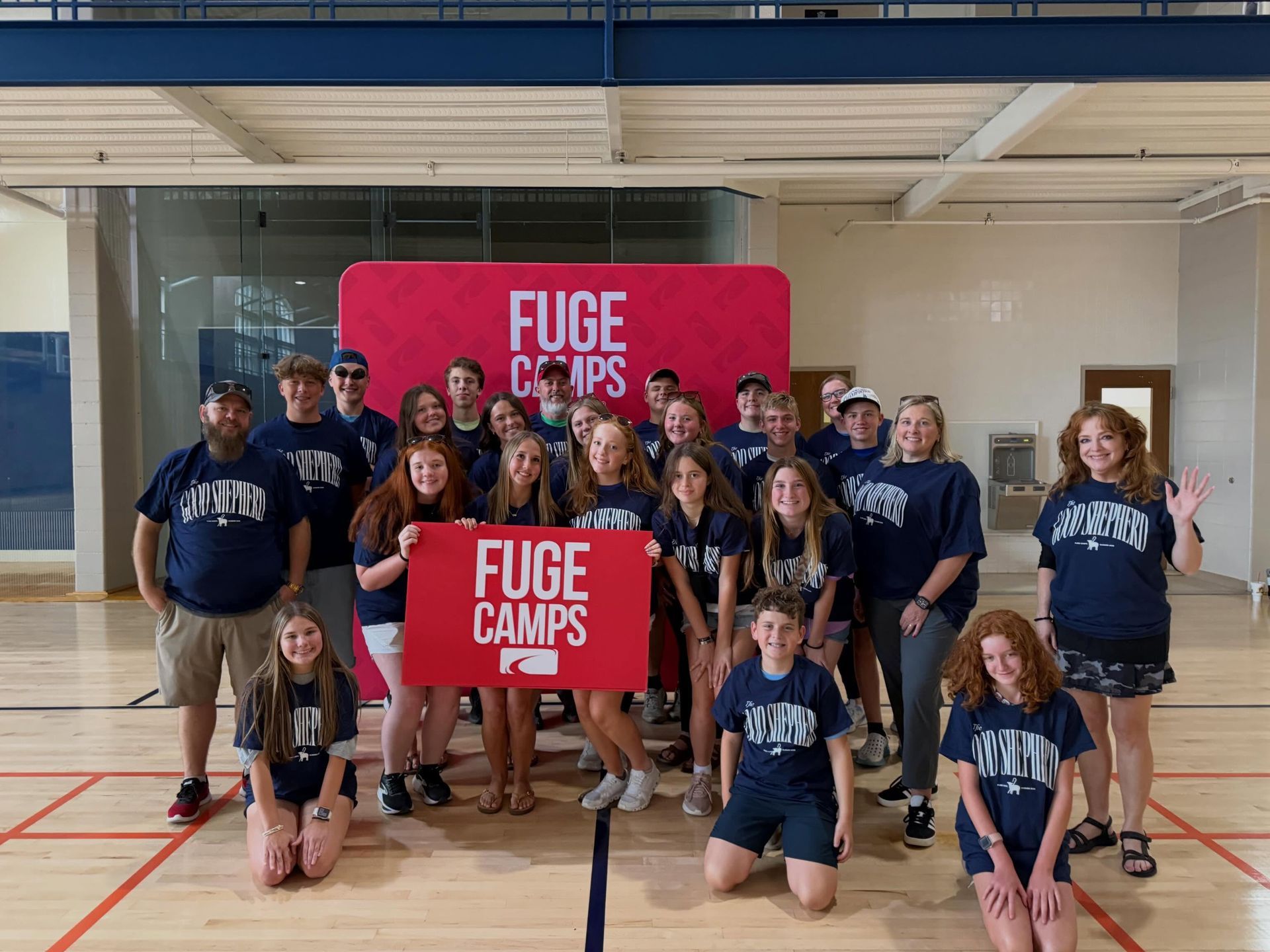 Group posing in a gym with two red FUCE CAMS banners, many wearing blue shirts.