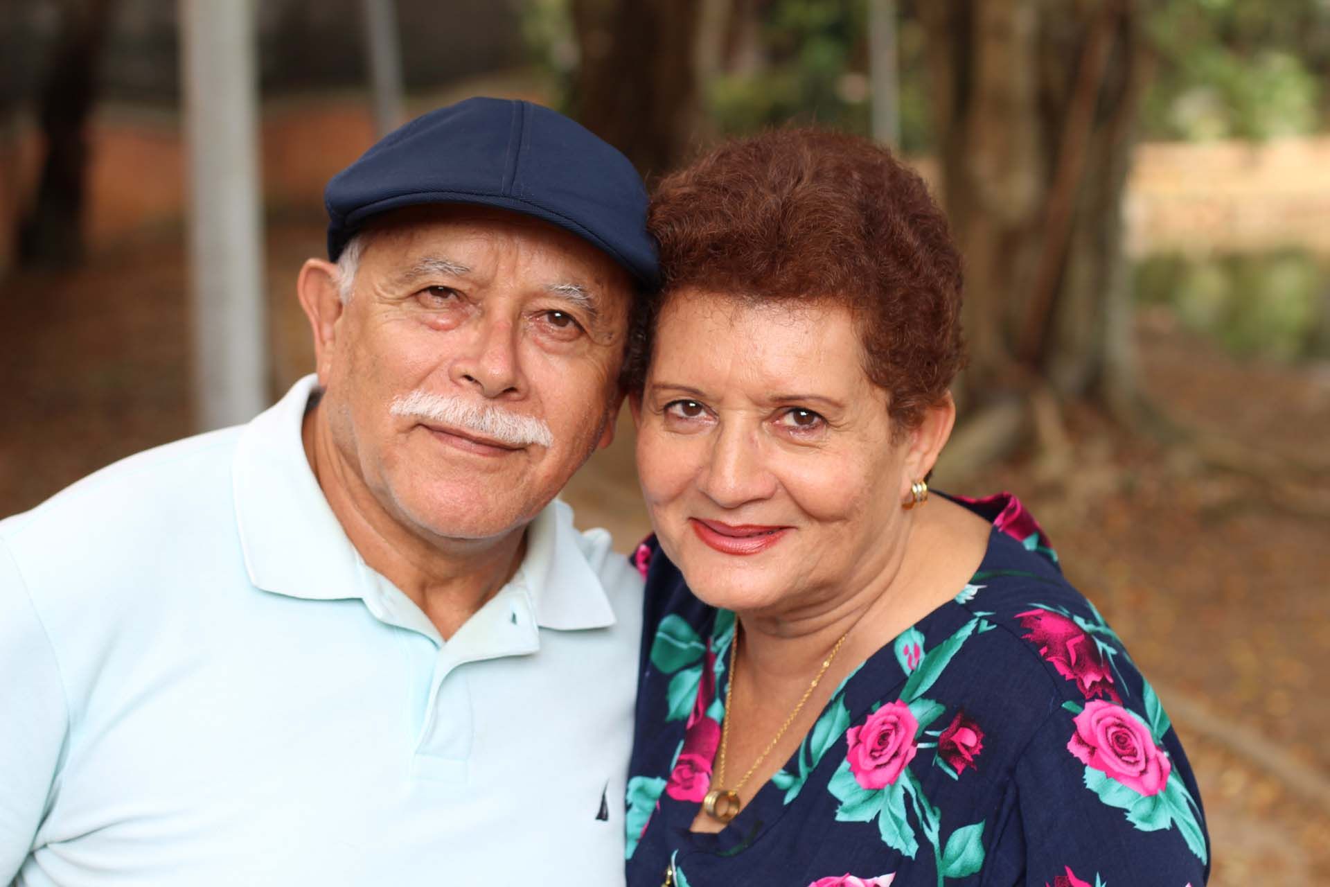 Two people standing close together outdoors, smiling at the camera, with trees in the background.
