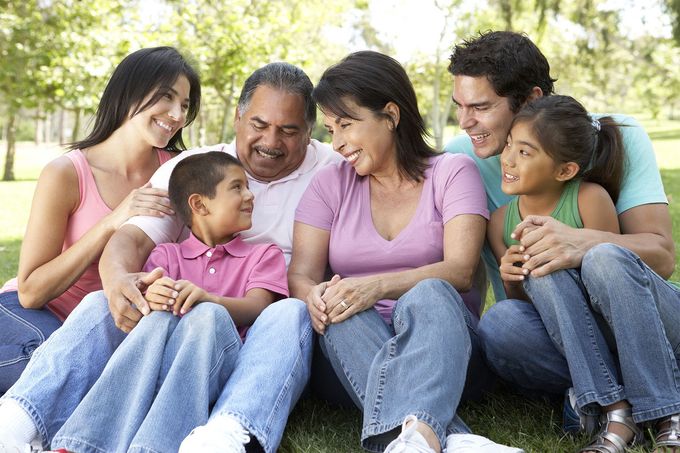 Family sitting together outdoors on a blanket, smiling and chatting in a park.