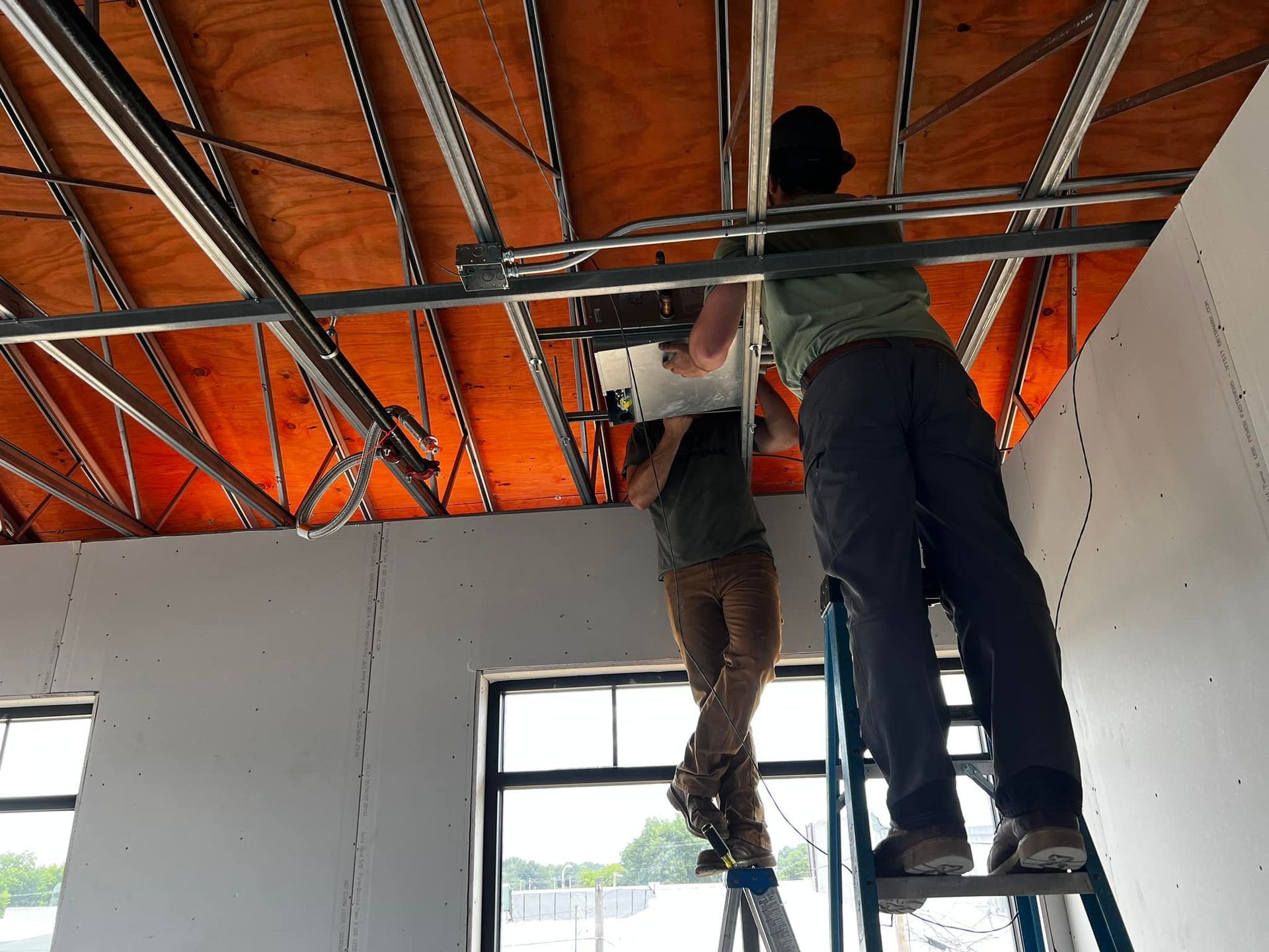 Two people installing something on a metal ceiling grid. One is on a ladder, the other is holding a square object.