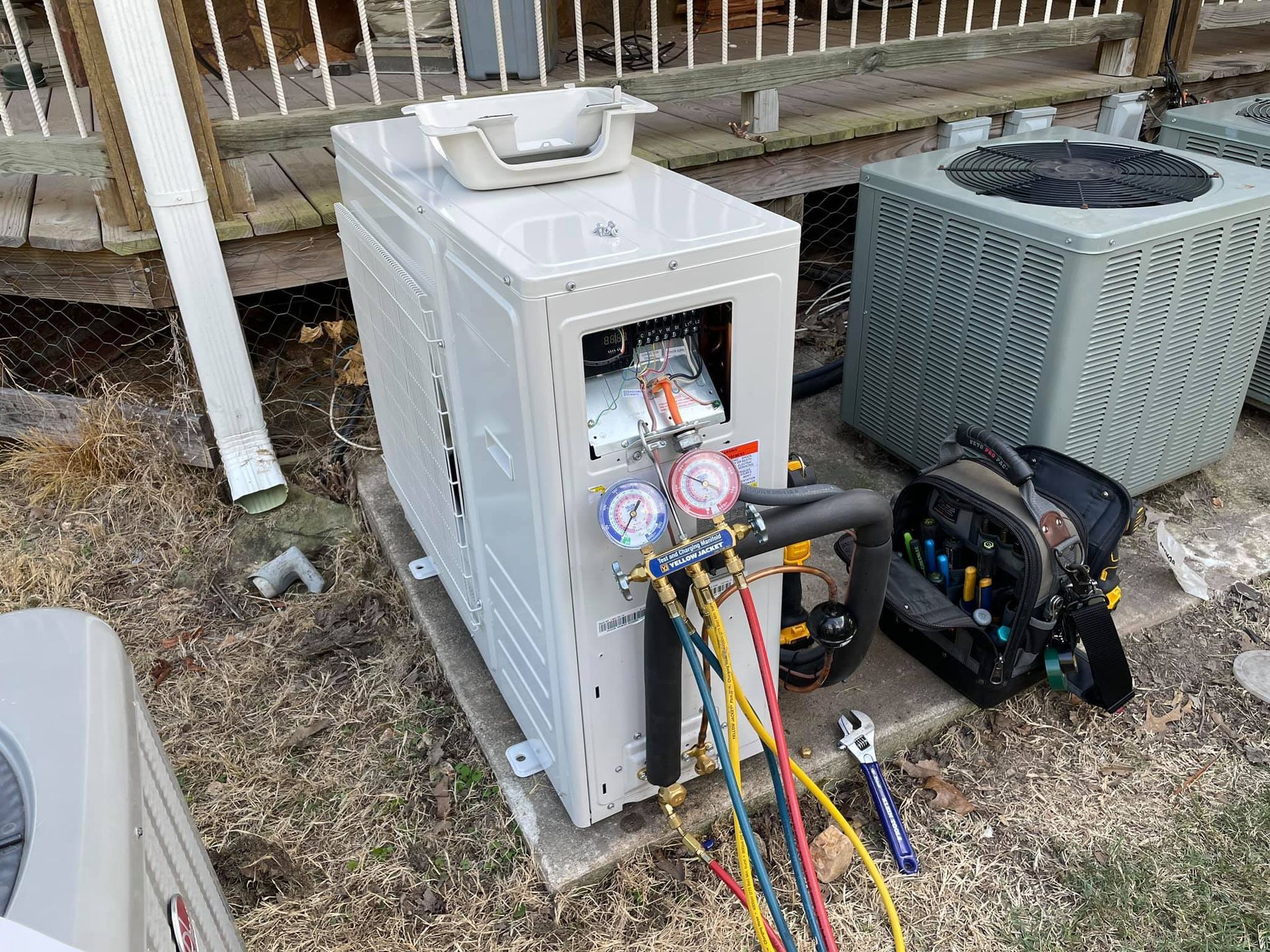HVAC unit being serviced outdoors with gauges connected. Various tools and another unit are nearby on a concrete slab.