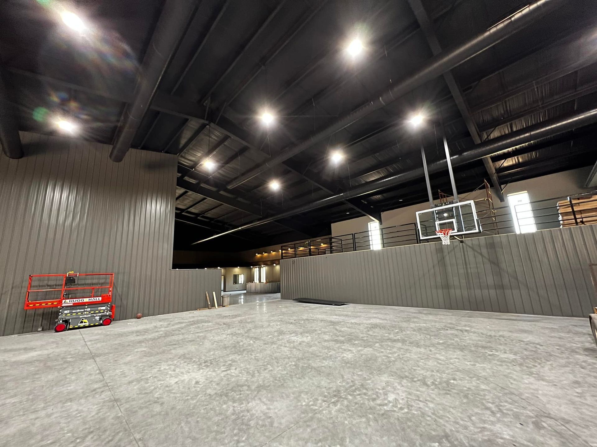 Indoor sports facility with a basketball hoop, gray walls, and overhead lighting. A lift is stationed by a wall.