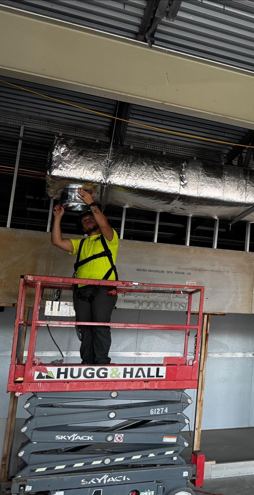 Construction worker on a lift installing material on a ceiling. The worker wears a safety harness and neon vest.