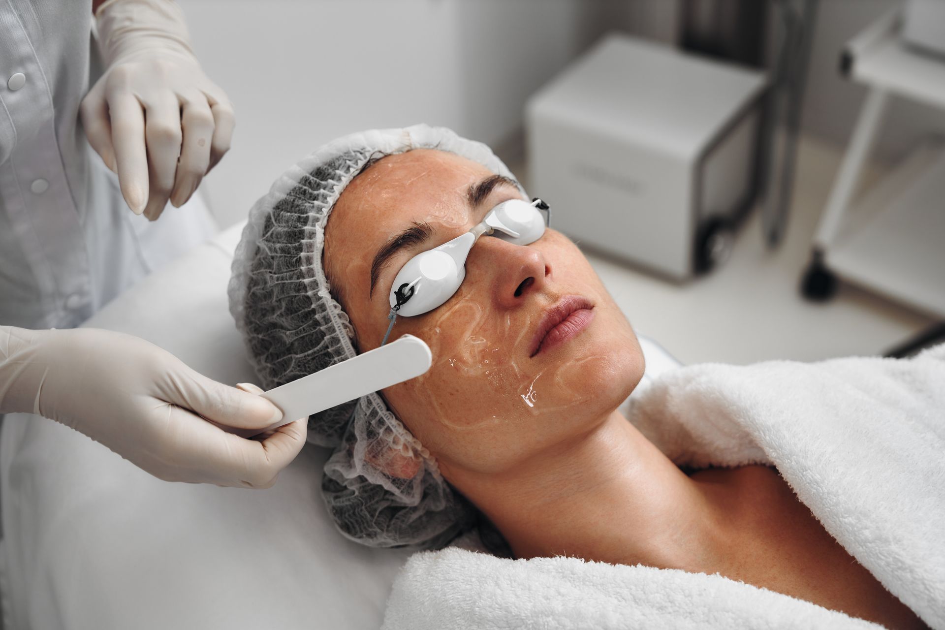 Esthetician applying facial treatment to a client lying on a spa bed with eye pads and hair cap
