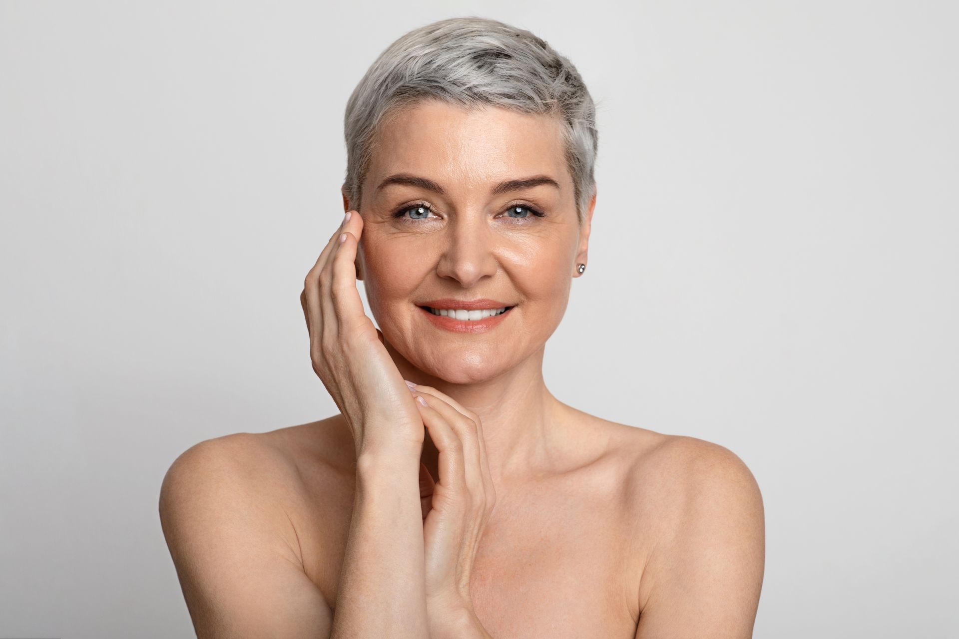 A woman with short gray hair is smiling and touching her face.