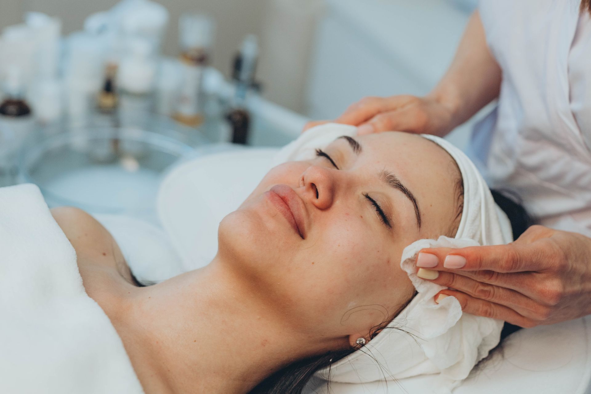 Person receiving facial at spa, eyes closed, being cleansed with a cotton pad.
