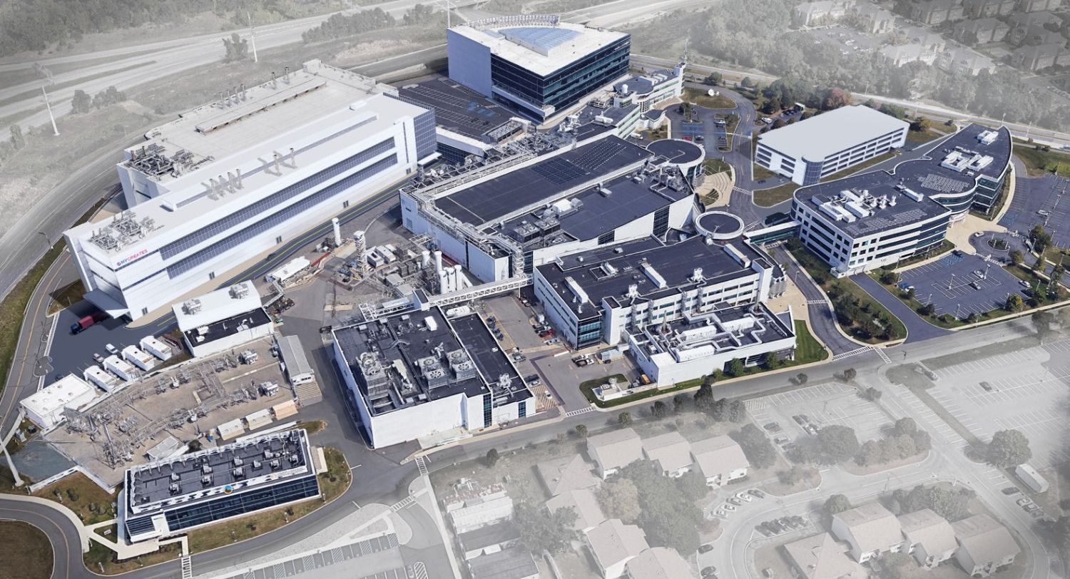 Aerial view of a large modern building complex with roads, parking lots, and surrounding terrain.