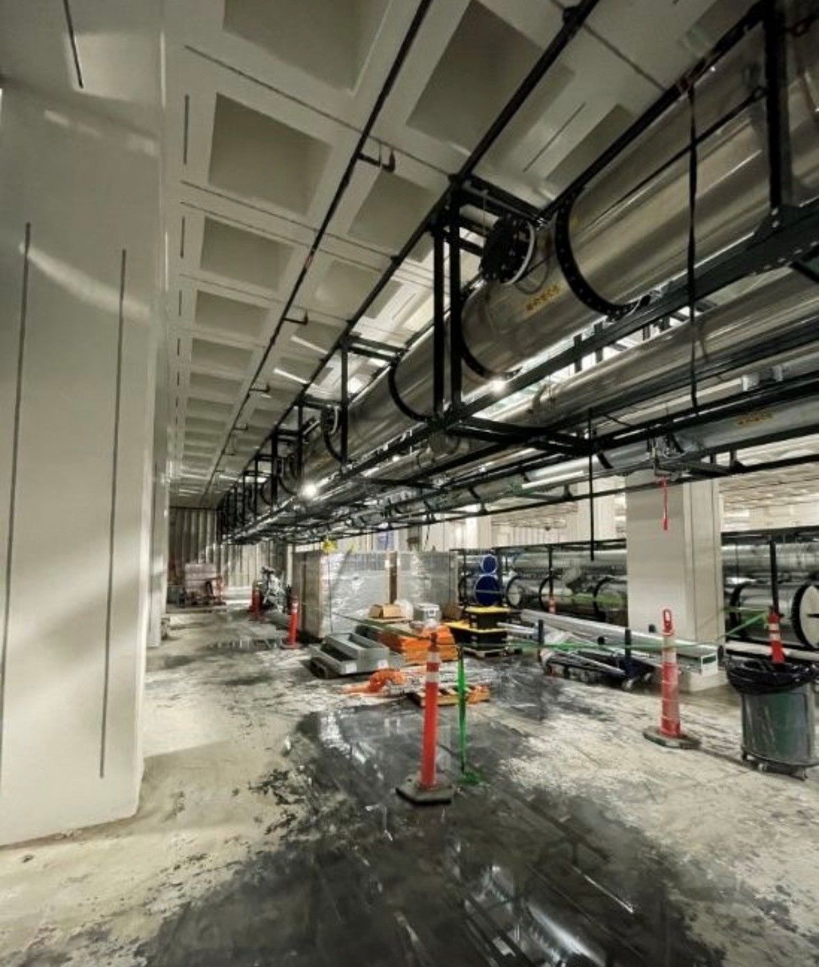 Construction site with exposed pipes, ducting, and orange safety cones in a long hallway