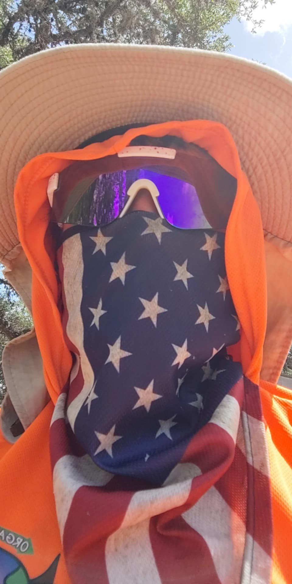 Person wearing a hat, sunglasses, orange hoodie, and a face covering with the American flag design.