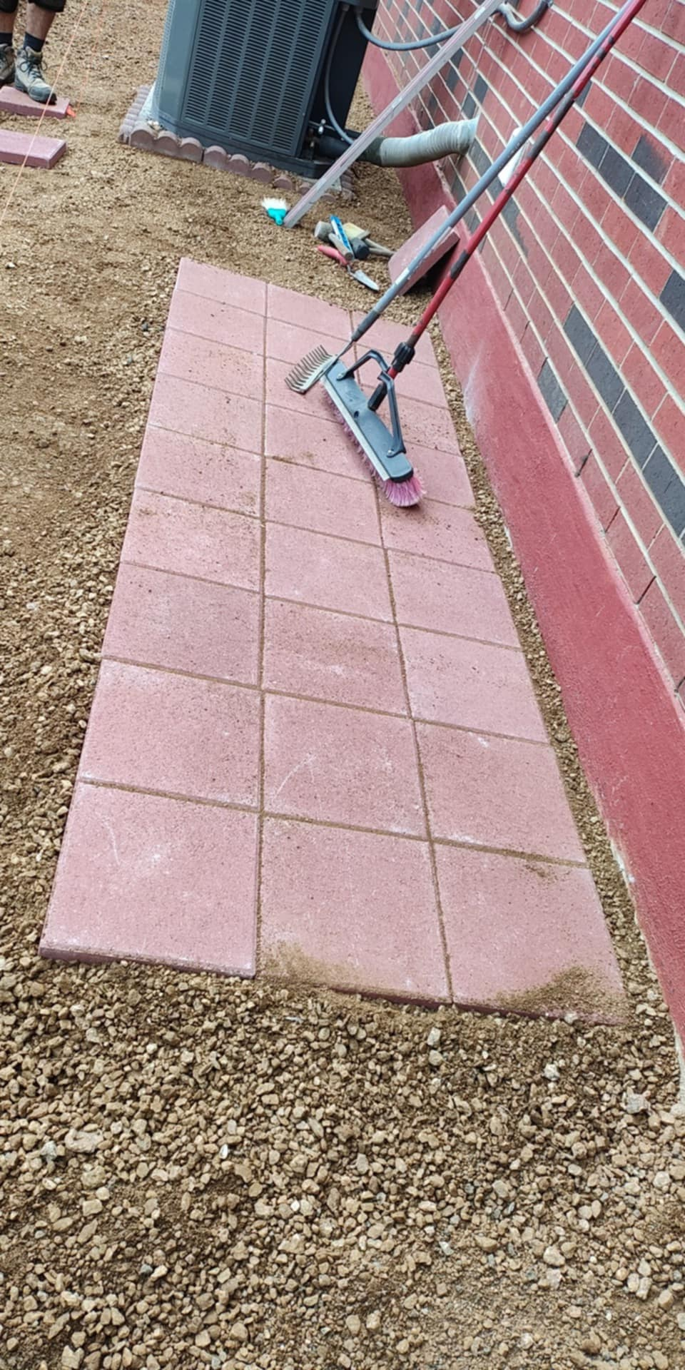 Red pavers are set near a building with a red brick wall. A tool sits on the pavers.