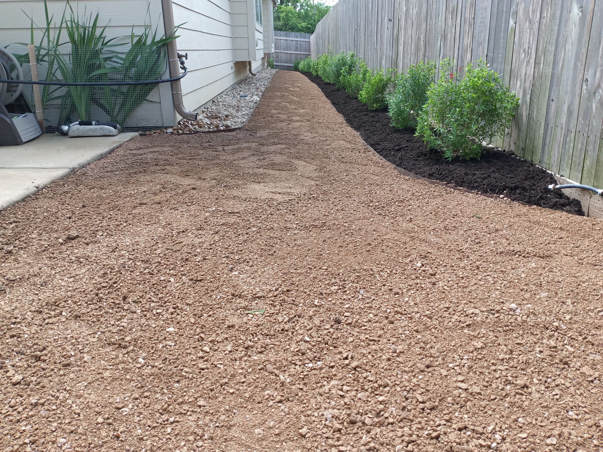 Wood-mulched pathway between a house and fence, with green shrubs along the fence and a concrete walkway on the left.