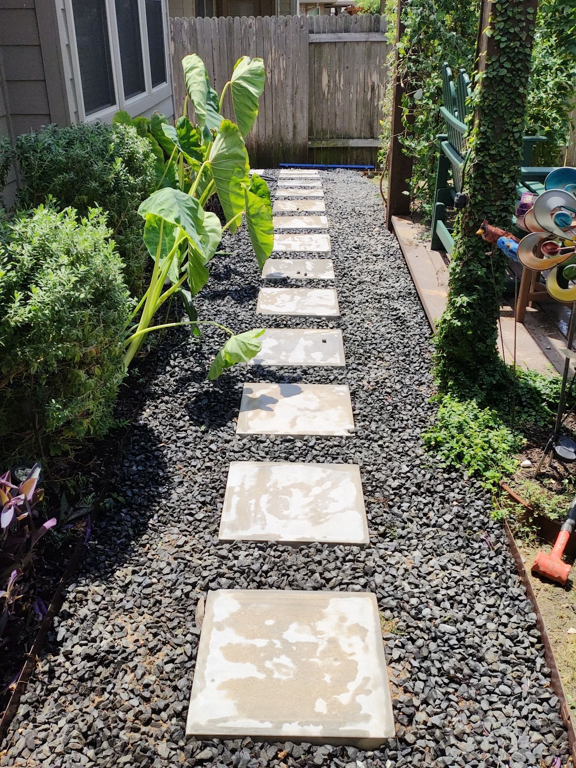 Stone path through a garden, bordered by black pebbles and green plants.