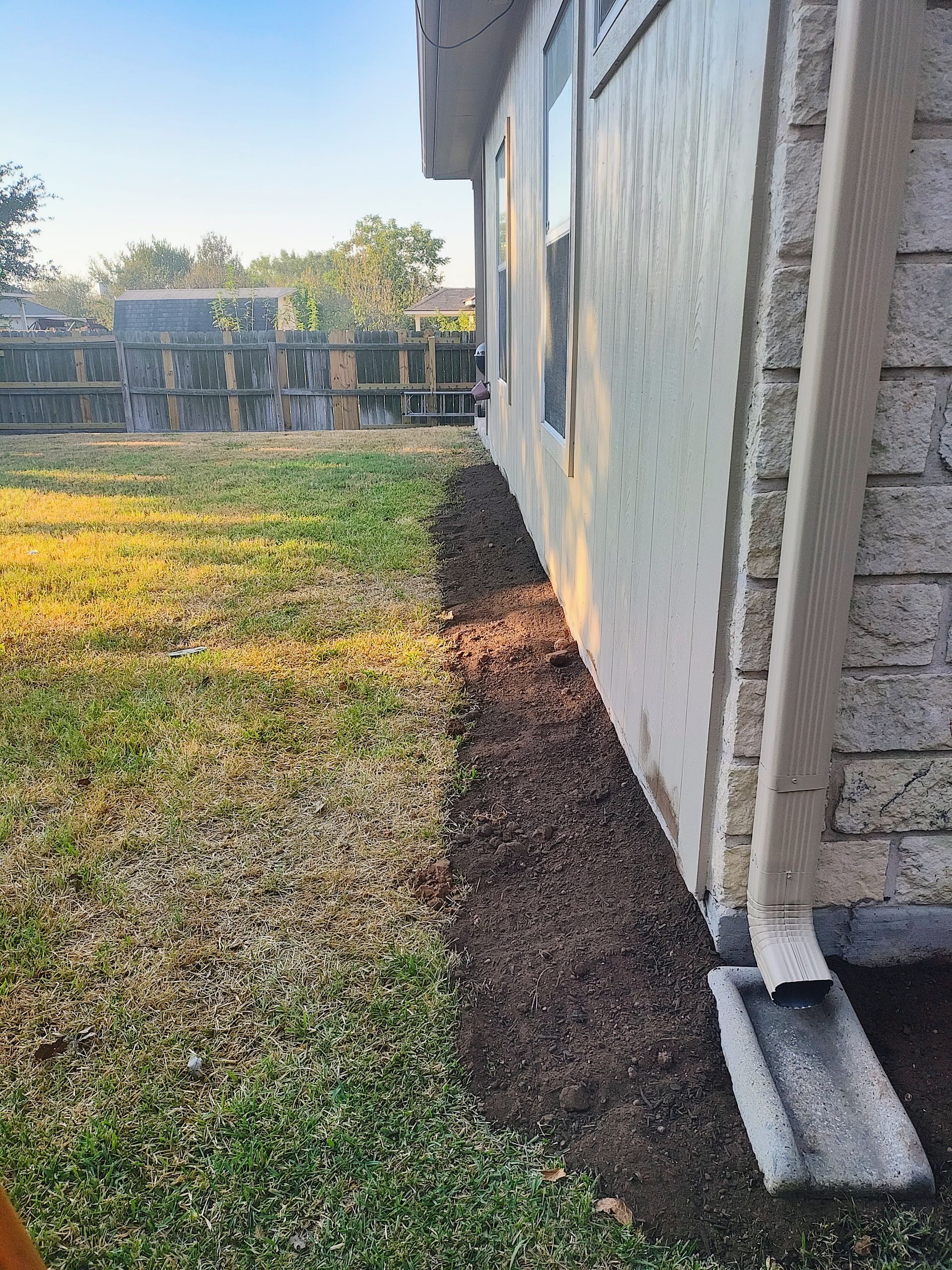 Gravel bed along exterior wall of house. Green grass and concrete foundation.