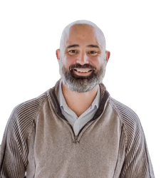 Bald man with gray beard smiles, wearing a light-colored sweater over a collared shirt.