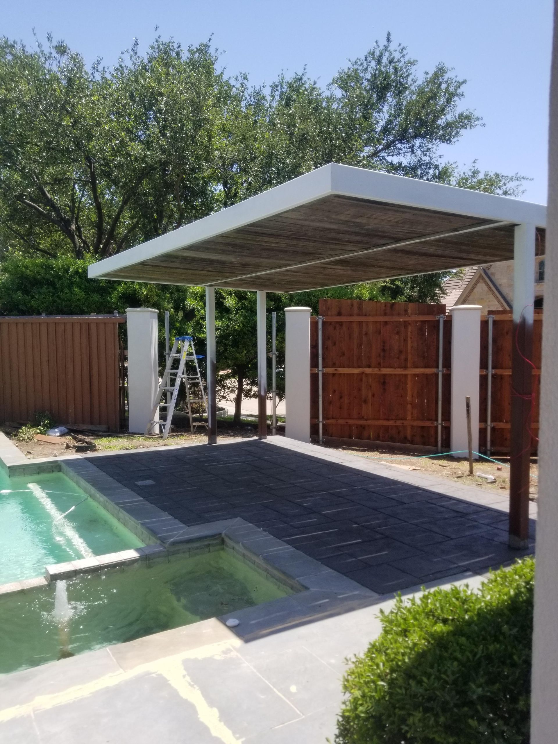 Backyard pool with a covered patio, wooden fence, and paved deck in bright daylight