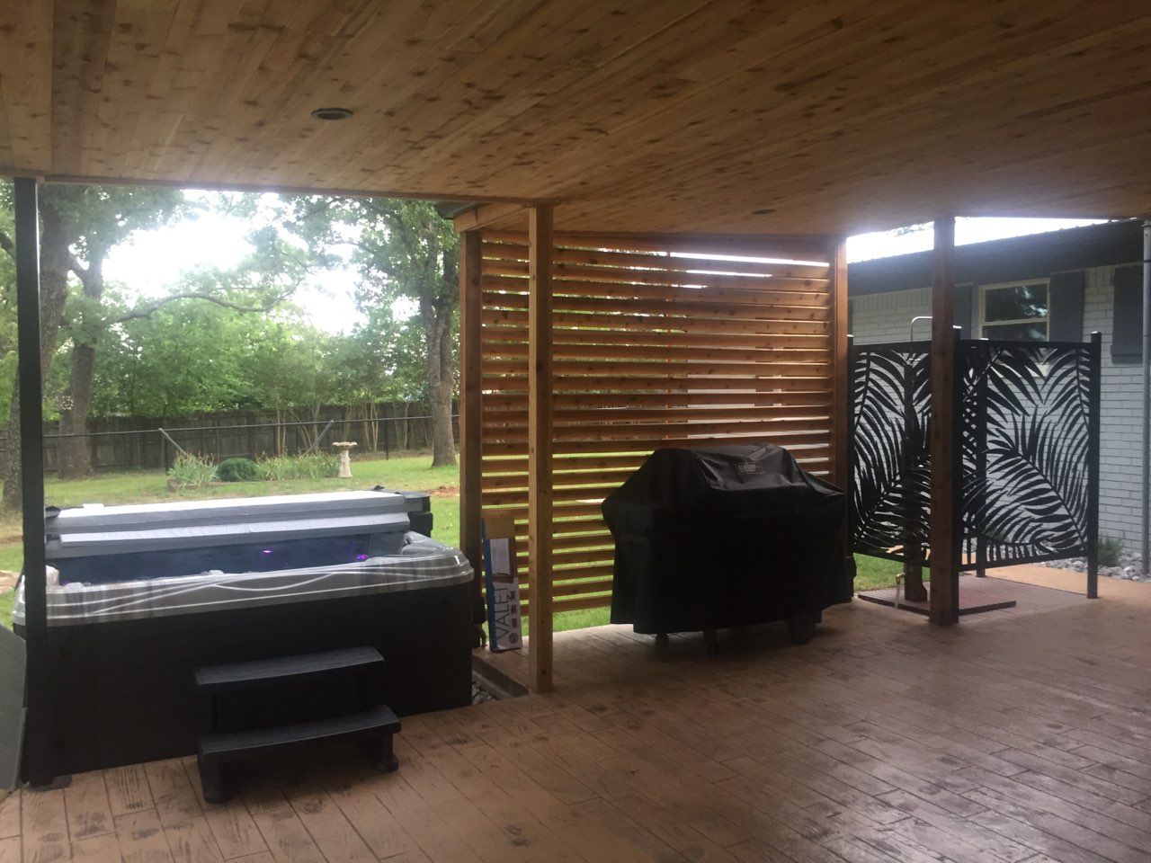 Covered patio with two black grills, wooden slat walls, and a view of a green yard beyond.