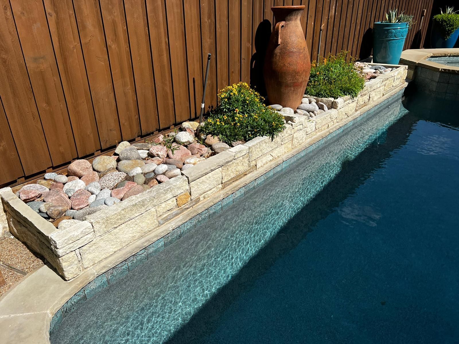 Backyard pool edge with blue water, stone border, wooden fence, and large clay pots.