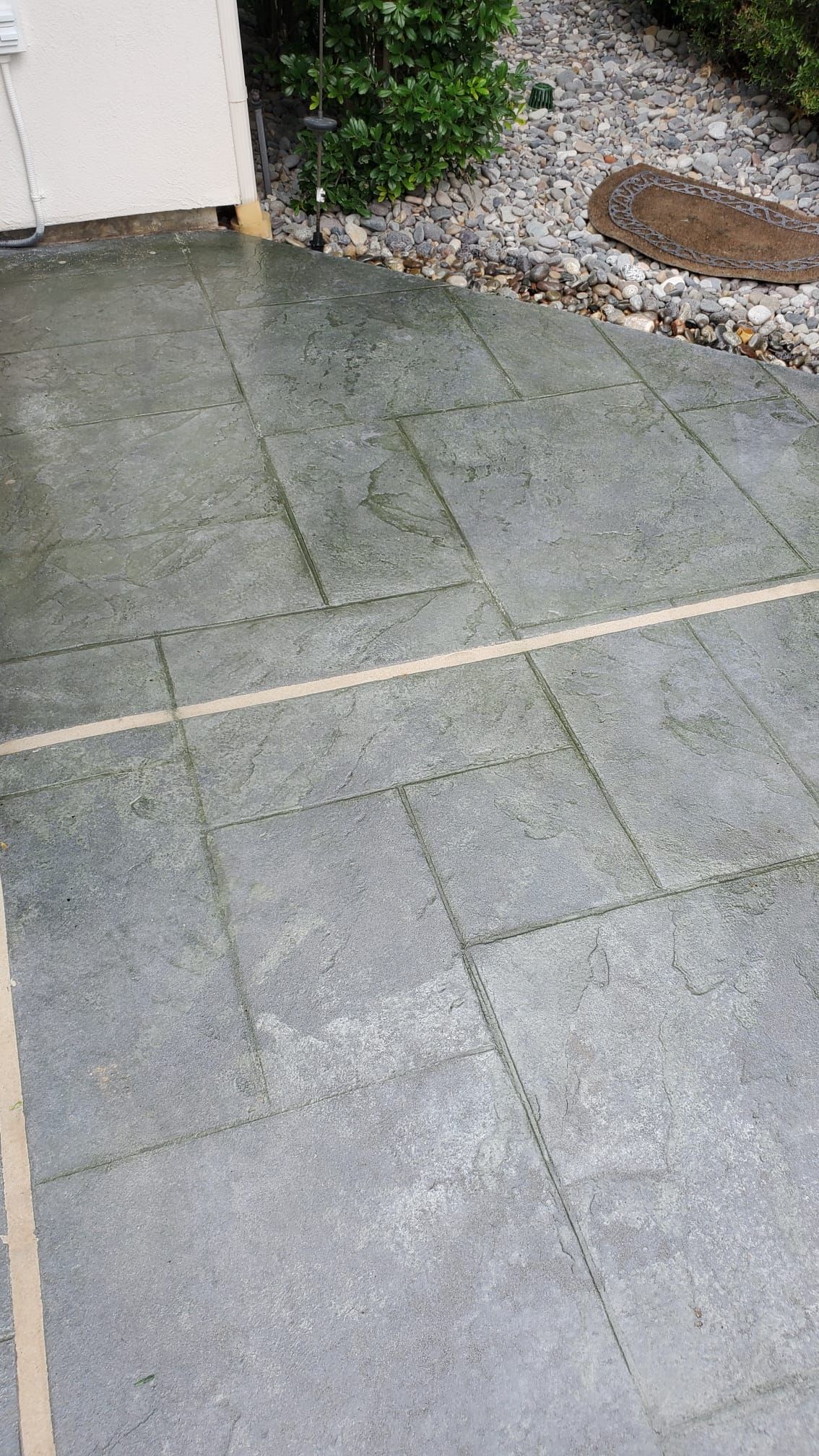 Wet gray patio tiles beside a gravel garden and house wall, with a narrow light-colored strip across the ground