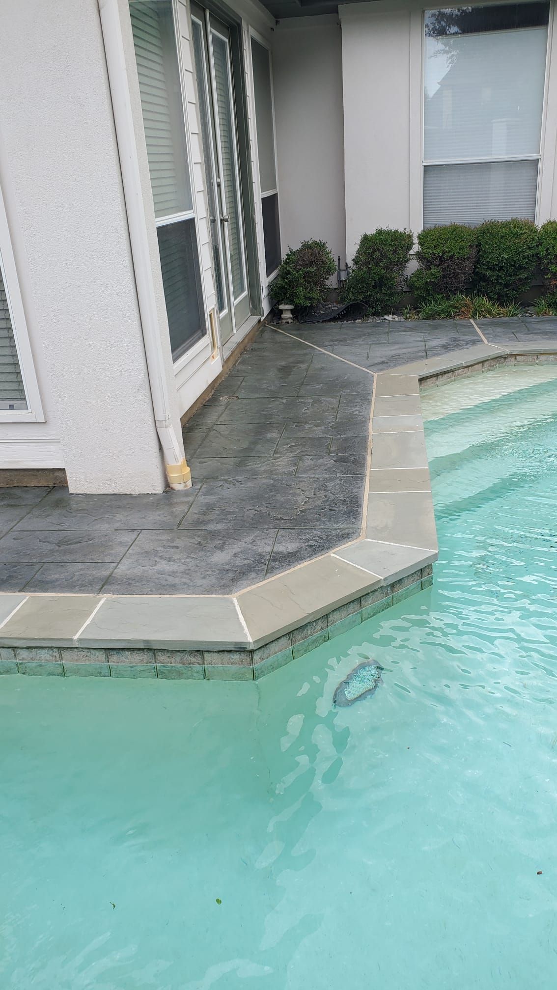 Poolside patio with gray stone coping beside turquoise water and white house exterior