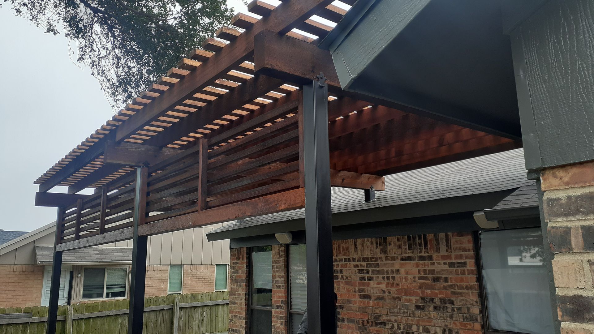 Covered driveway/carport with dark posts, brick walls, and a corrugated roof beside a house.
