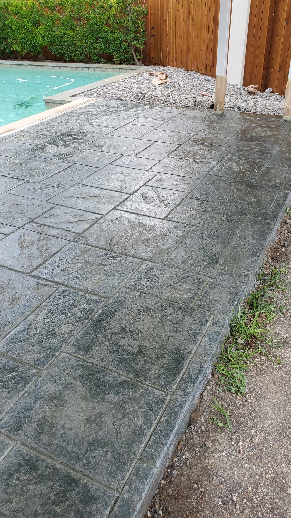 Wet stone patio beside a pool and wooden fence, with a gravel edge and white column