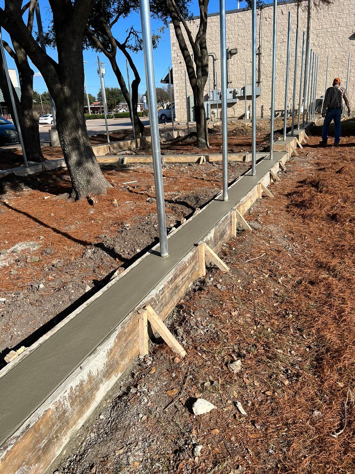 Concrete retaining wall along a dirt path with metal posts, trees, and a building in the background