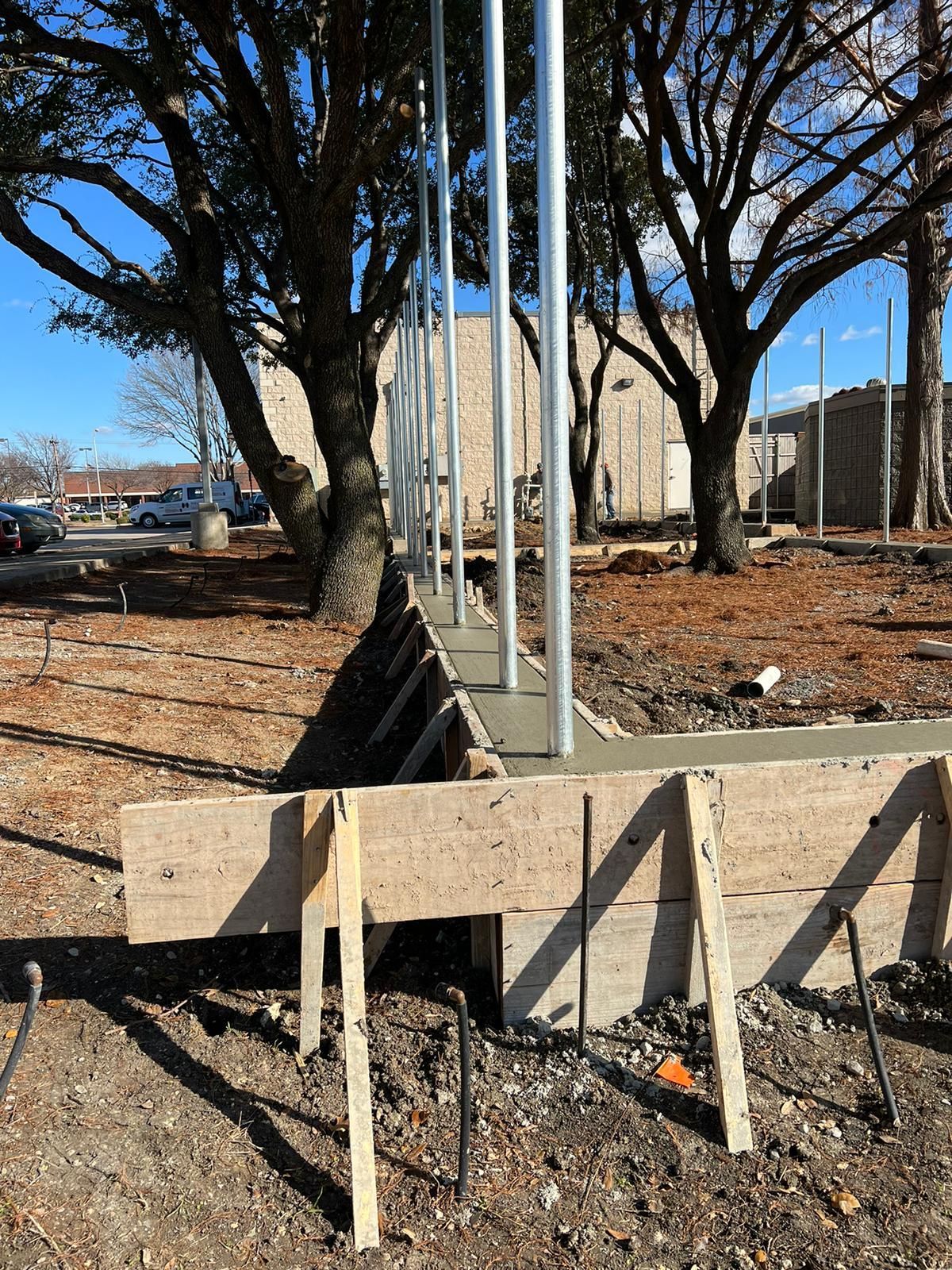 Concrete foundation with vertical steel poles and wooden stakes at a construction site under bare trees.