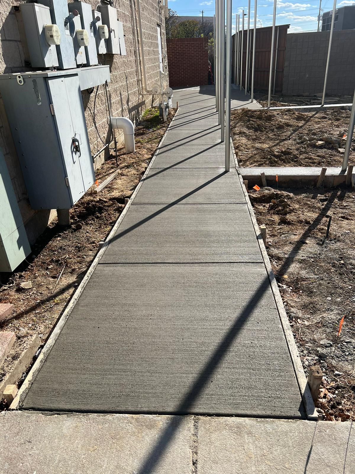 Concrete walkway between utility meters and a stone wall, bordered by dirt and strong sunlight.