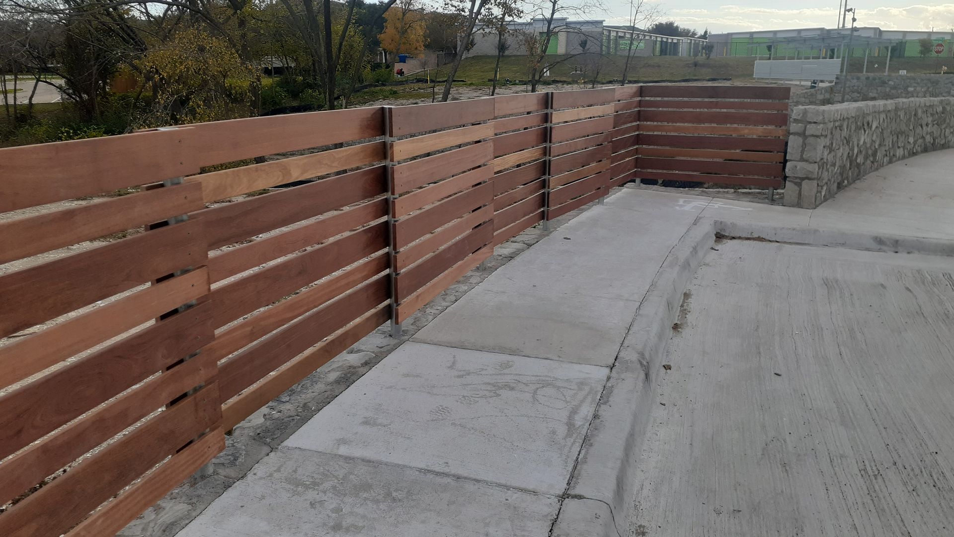 Wooden fence beside a concrete walkway and road, with a stone wall at the end.