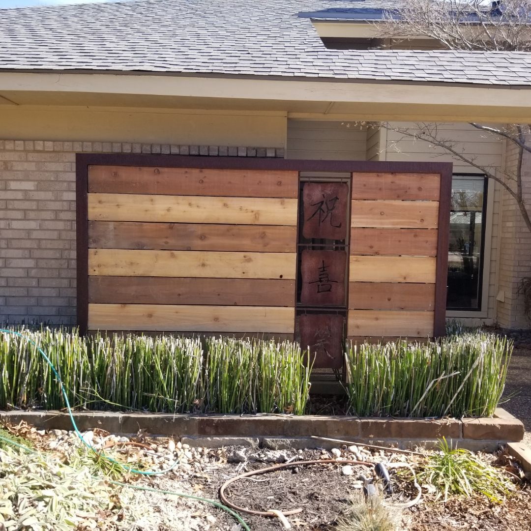 Wooden shed-like structure against a brick house, with a dark door and a row of green plants in front.
