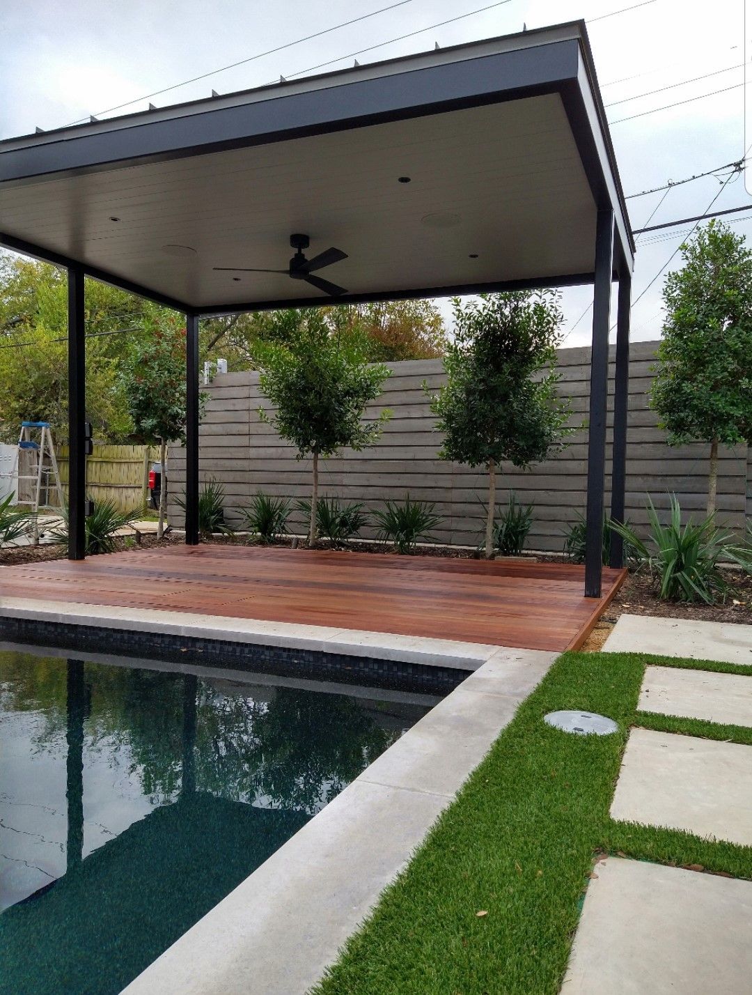 Modern backyard pool with a covered patio, wooden deck, and green lawn stepping stones