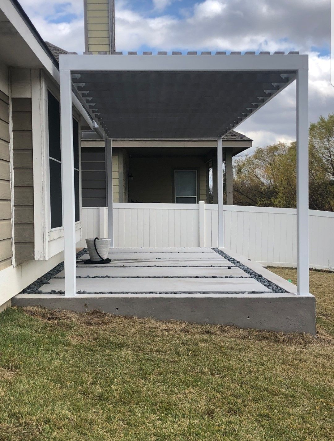 Covered backyard patio with white posts, deck framing, and a white fence beside a house.