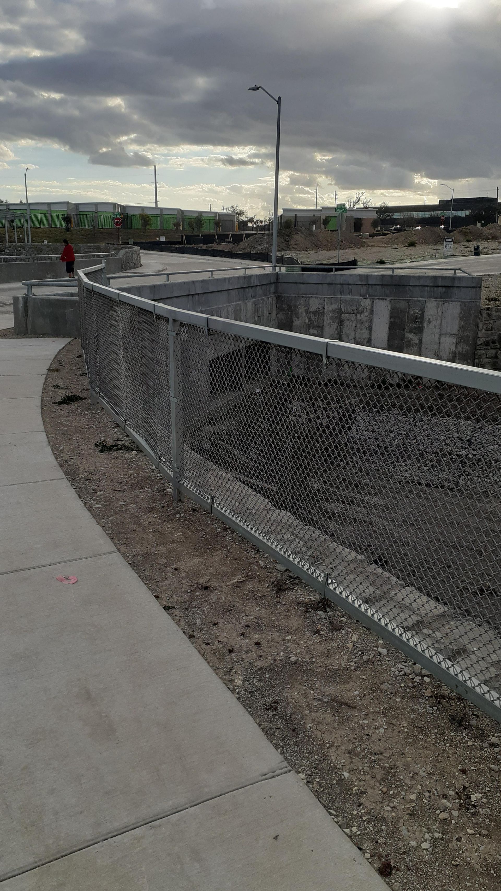 Curved sidewalk beside a chain-link fence overlooking a cloudy waterfront and distant buildings.