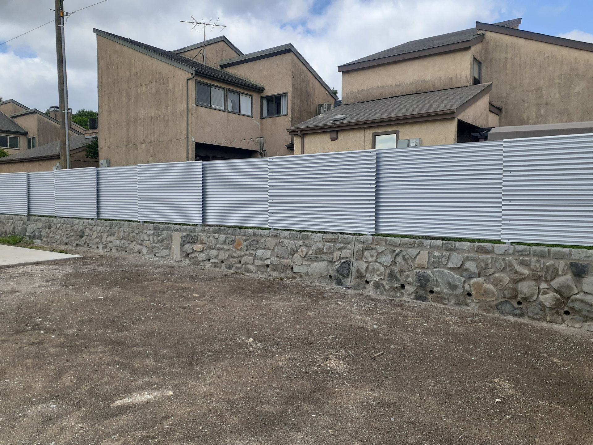 Rural residential houses behind a gray corrugated fence and stone wall under an overcast sky