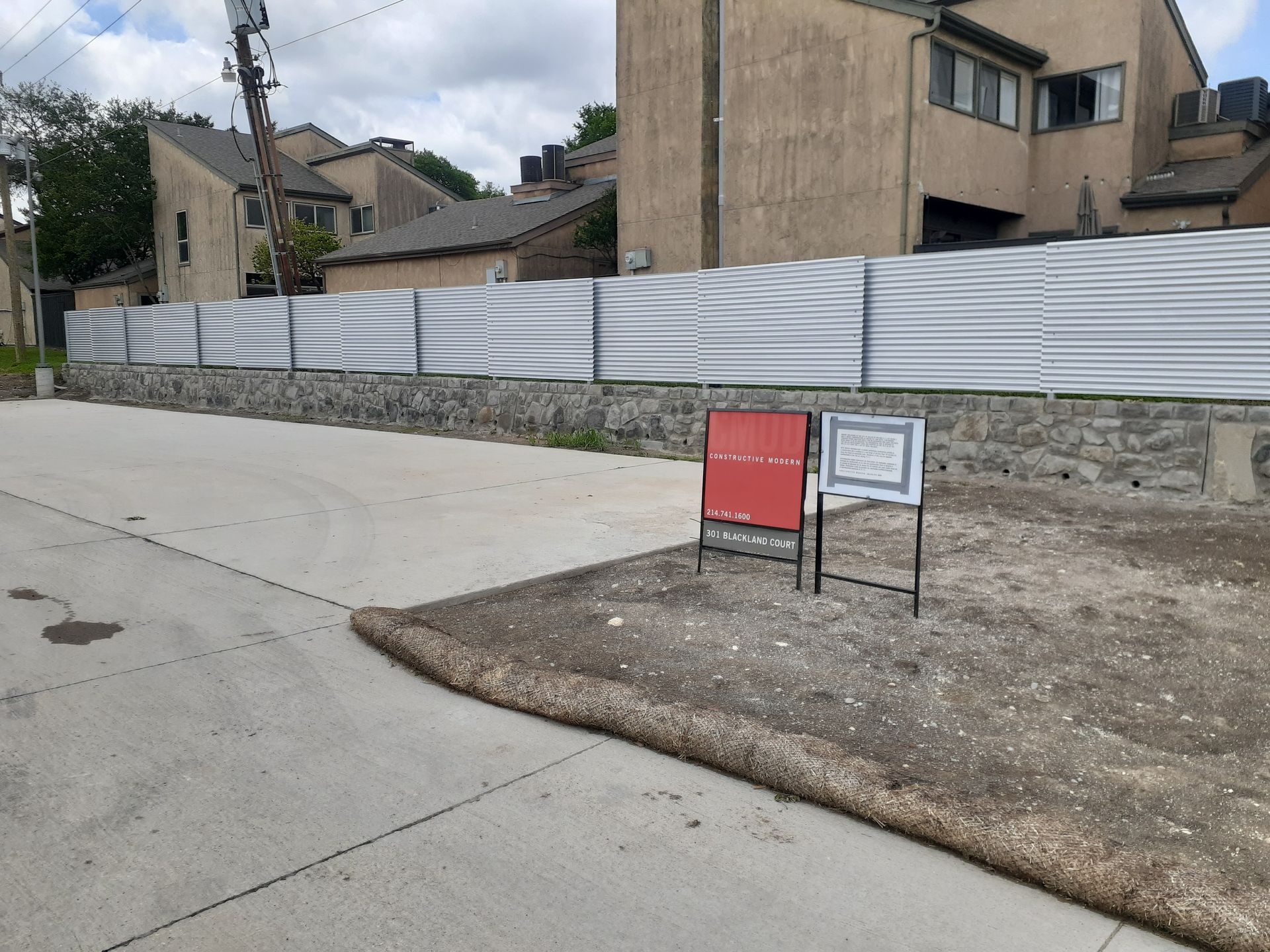 Vacant lot beside a sidewalk with a red sign and white fence in front of tan buildings