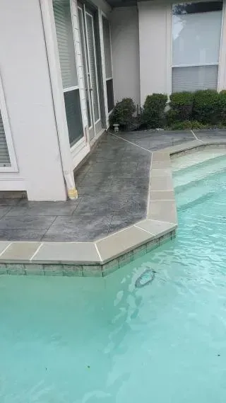 Poolside patio with light gray stone coping and turquoise water beside a white building.