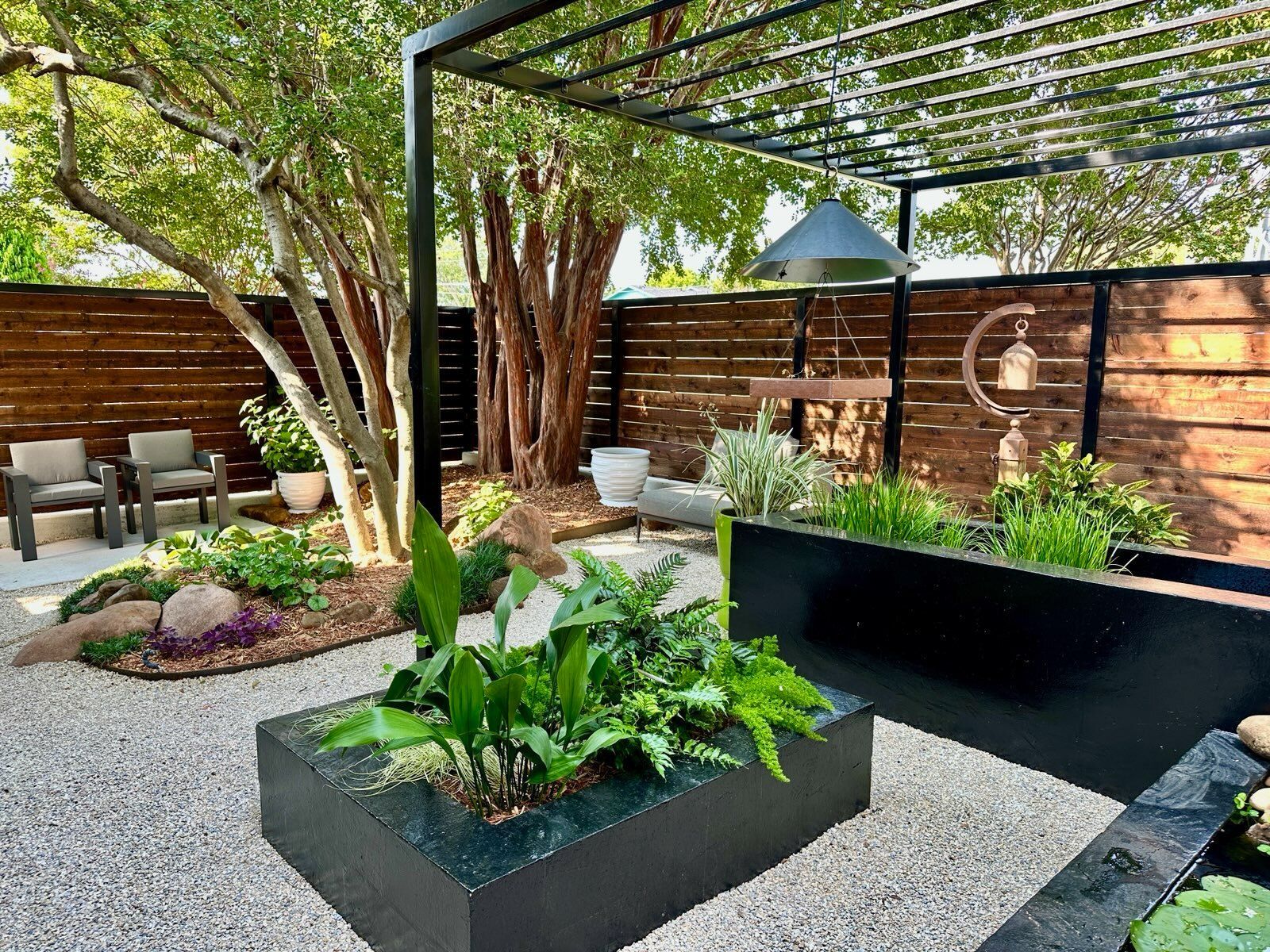 Modern backyard patio with raised black planters, trees, seating, and a pergola overhead