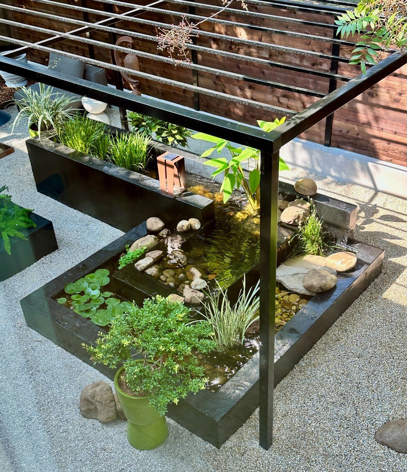 Small outdoor water garden with black stone planters, rocks, and green plants beside a railing.