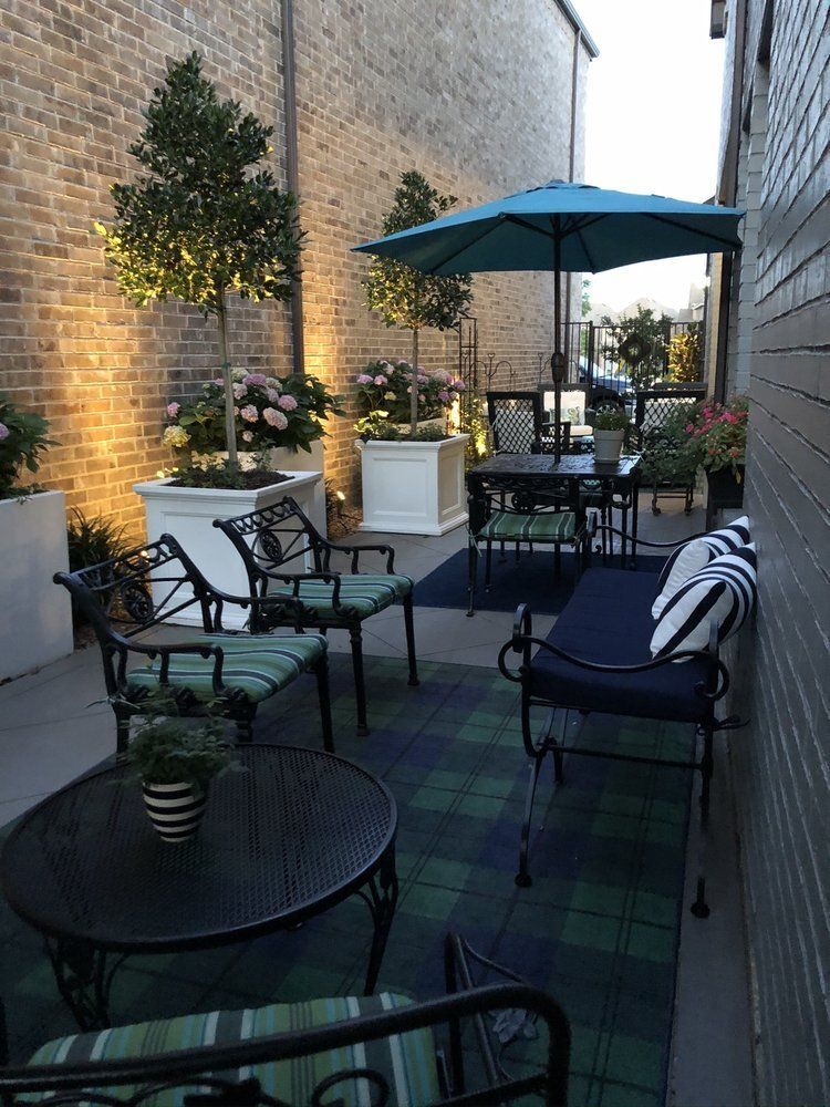 Small outdoor patio with chairs, round tables, blue umbrella, plants, and string-lit brick walls.